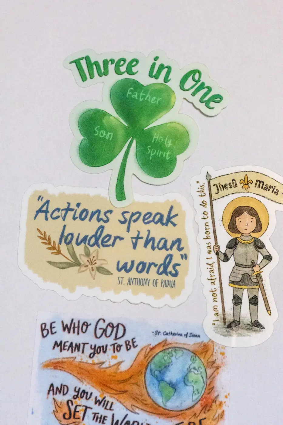 Original saint stickers — a Trinity shamrock, St. Anthony's 'actions speak louder than words,' St. Joan of Arc, and St. Catherine of Siena